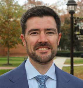 Headshot of Cedar Riener. A white man with short black hair and a short trimmed beard, wearing a blue jacket, blue tie, light blue shirt. 